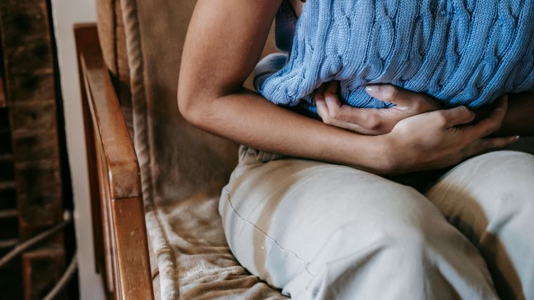 Image of a woman in a blue vest sat on a sofa while wrapping her arms around her stomach in pain.